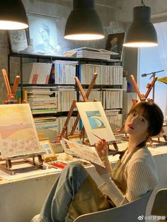 a woman sitting in front of an easel with paintings on it and two lamps hanging from the ceiling