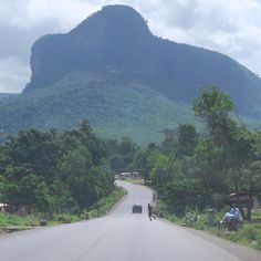 two people riding motorcycles down the middle of a road in front of a large mountain