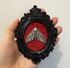 a hand holding a small black frame with a silver moth on it's side