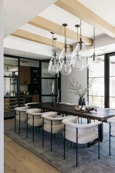 a dining room table with chairs and lights hanging from it's ceiling, in front of large windows