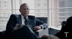 a man in a suit and tie sitting on a chair next to a window looking at something