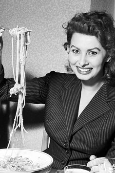 a woman sitting at a table with noodles in front of her and holding a fork