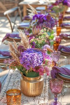 the table is set with purple flowers and gold vases on it, along with other place settings