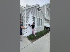 two people are decorating a white christmas tree on the side of a gray house