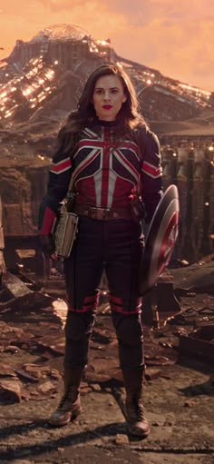 a woman dressed as captain america stands in front of a destroyed area with mountains behind her