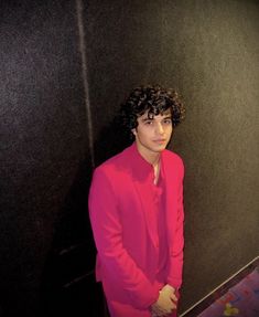 a man in a pink suit standing next to a black wall with his hands on his hips