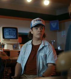 a young man sitting in front of a computer monitor wearing a hat and looking at the camera