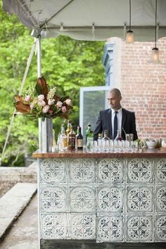 a man standing behind a bar with bottles and glasses on it, in front of a brick building