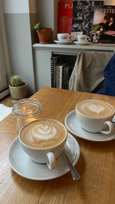 two cups of cappuccino sit on a wooden table in front of a window