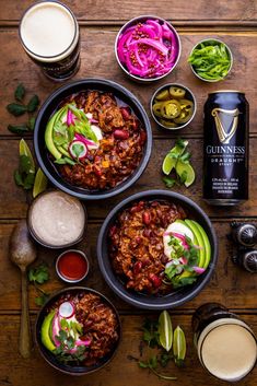 three bowls filled with chili and veggies next to two cans of beer