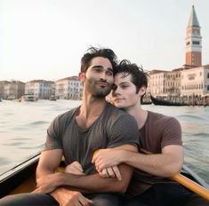 two young men are sitting in a boat on the water near some buildings and one is holding his arm around the other man's shoulder