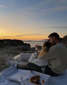 a man and woman sitting on the beach at sunset with pizza in front of them