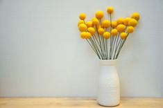 a white vase filled with yellow flowers sitting on top of a wooden table next to a wall