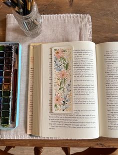 an open book sitting on top of a wooden table next to a paintbrush and watercolor