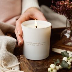 a person holding a white candle on top of a wooden table next to some flowers