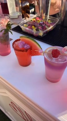 three drinks are sitting on a table with flowers in the vases and one has a slice of watermelon