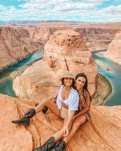 two women sitting on the edge of a cliff in front of a river and canyon