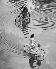 black and white photograph of two people riding bikes in water with ripples on the ground