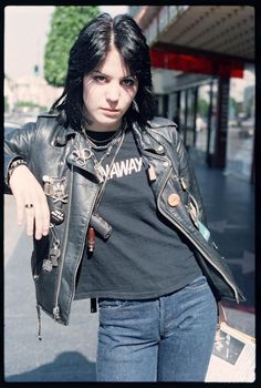 a woman with black hair wearing a leather jacket and holding a skateboard in her hand