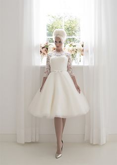 a woman standing in front of a window wearing a short white dress with lace sleeves