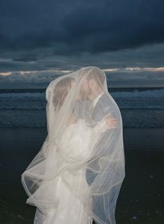 a bride and groom kissing under a veil on the beach at night with dark clouds in the background