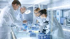 three people in white lab coats looking at something on a table with microscopes and test tubes