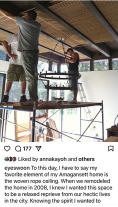 two men standing on top of a wooden beam in a room next to a window