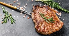 a piece of steak on a black surface with rosemary sprigs and seasoning