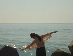 a woman standing on rocks near the ocean with her arms spread out to catch something