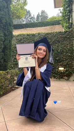 a woman in graduation gown sitting on the ground holding up her diploma and smiling at the camera