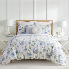 a white bed with blue and green flowers on the comforter, pillows and lamps