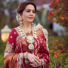 a woman in a red and gold sari with jewelry on her neck, standing outside