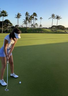 a woman in white shirt and skirt playing golf on green with palm trees behind her