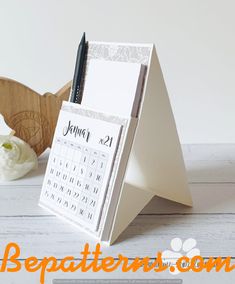 a desk calendar sitting on top of a wooden stand next to a pen and flower