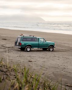 a green pick up truck parked on the beach next to the ocean and grass in front of it