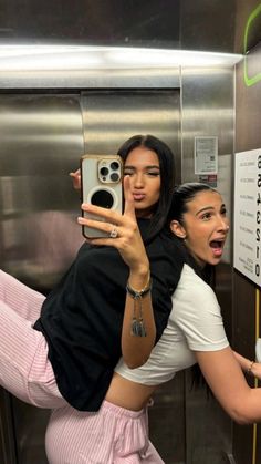 two women taking selfies in an elevator while one takes a photo with her phone