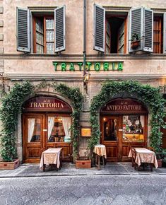 the outside of a restaurant with two tables and chairs in front of it, surrounded by greenery