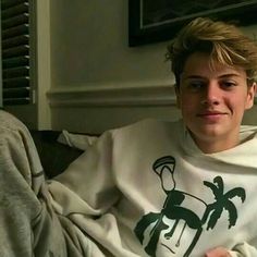 a young man sitting on top of a couch wearing a white shirt with palm trees on it