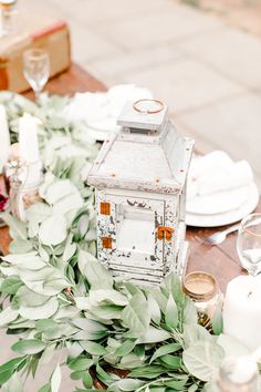 a wooden table topped with lots of greenery next to white plates and silverware