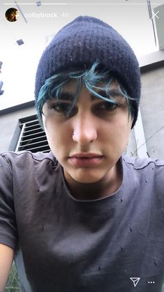 a young man with blue hair wearing a beanie and looking at the camera while standing in front of a building