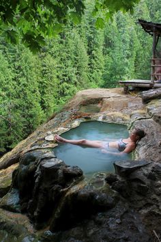 a woman laying in a pool of water next to some rocks and trees on the side of a cliff