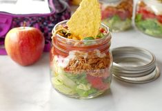 a jar filled with salad and chips next to an apple