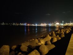 the city lights shine brightly in the distance over the water and rocks on the shore