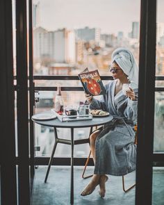 a woman sitting at a table with a book in her hand and looking out the window