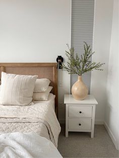 a bed with white sheets and pillows in a bedroom next to a nightstand with a vase on it