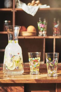 three glasses and a pitcher sitting on a table next to a shelf with other glassware