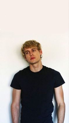 a young man standing in front of a white wall wearing a black t - shirt