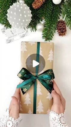 a person holding a present box in front of a christmas tree with pine cones on it