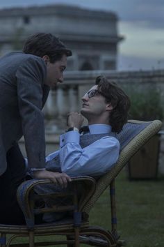 two men in suits are sitting on chairs and one is touching the other's neck