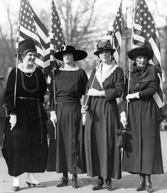 three women in long dresses and hats holding american flags on the street, with one woman standing behind them